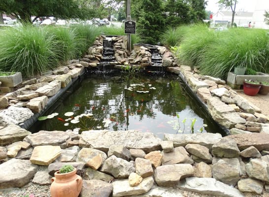 A serene garden pond with a small waterfall, surrounded by rocks and greenery.