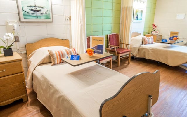 A shared bedroom with two beds, side tables, and seating area in a senior living facility