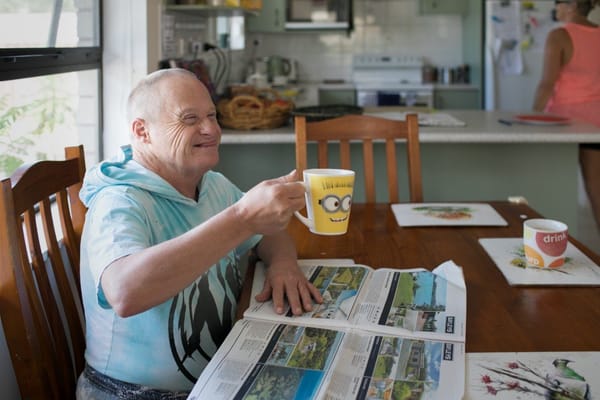 A resident holding a coffee mug, smiling at the dining table with newspapers spread out.