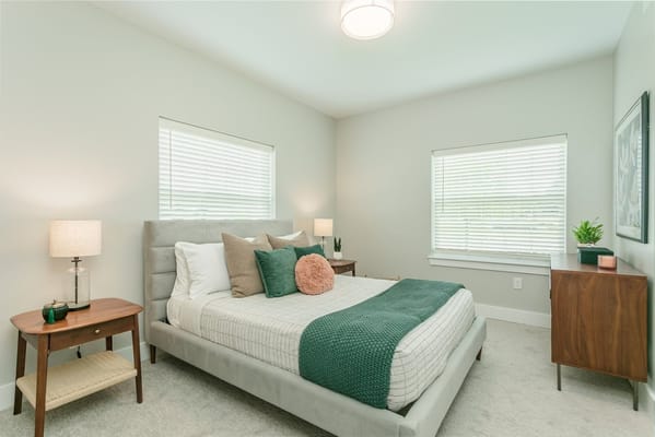 A well-decorated bedroom featuring a bed, bedside table, and natural light from two windows.