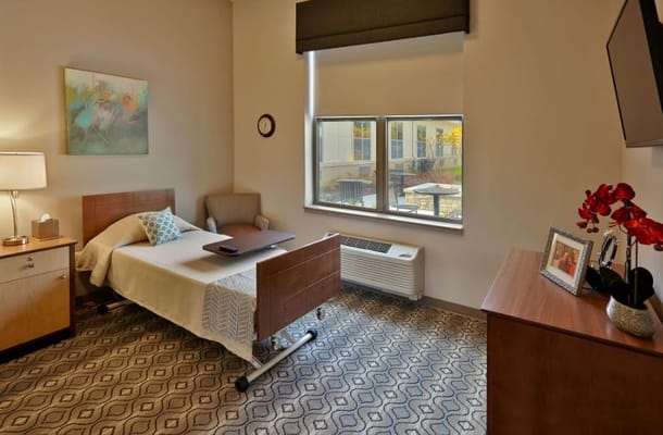 A cozy bedroom in Wellbrooke of South Bend with a bed, chair, and window overlooking an outdoor area.