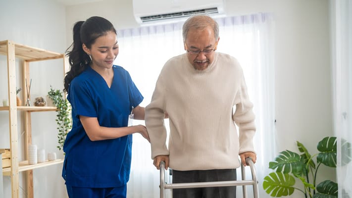 A caregiver assisting an elderly man with a walker in a bright room