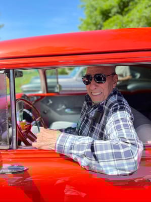 An elderly man in sunglasses smiling from a red car.
