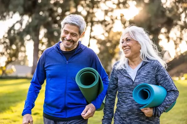 Two seniors smiling and walking with yoga mats in a park