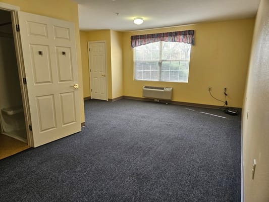 A vacant resident room with a window and carpeted floor
