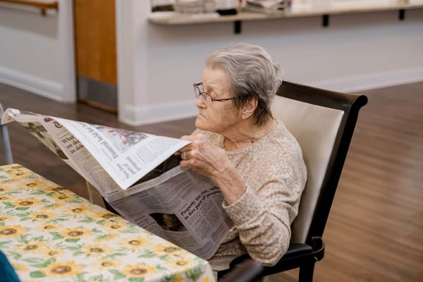 An elderly woman reading a newspaper at a table