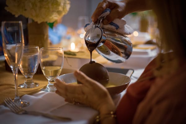 A hand pouring chocolate sauce over a dessert at a dining table