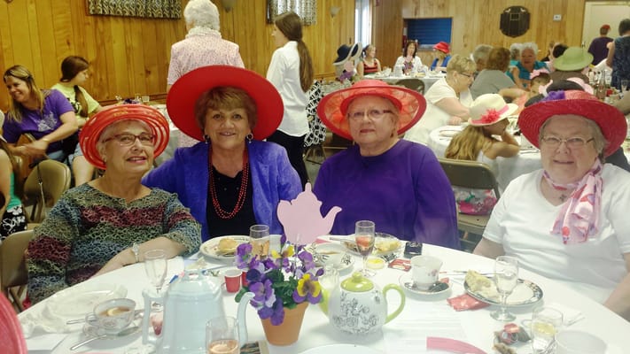 Four women in colorful hats enjoying tea together at a social event.