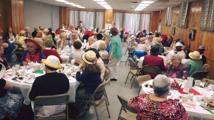 A vibrant social event at a senior living facility with many residents wearing hats and enjoying a meal together.