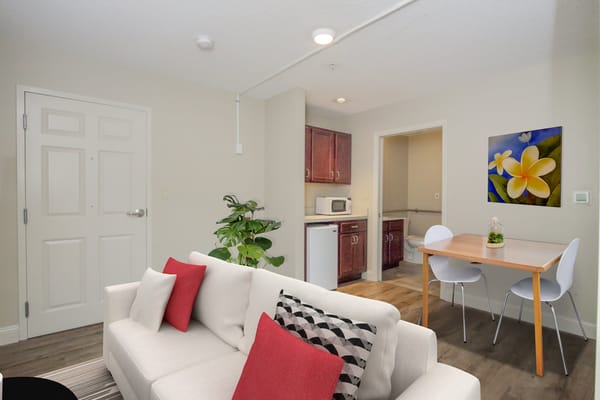 Living room with a sofa, dining area, and kitchenette in a senior living apartment.