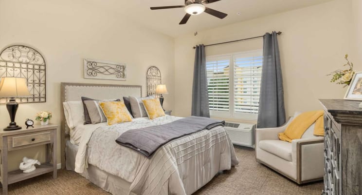 A furnished bedroom with a queen-sized bed and decorative pillows, featuring a chair and natural light from a window.