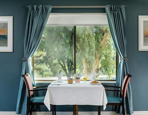 A dining table set near a large window with curtains, overlooking greenery.