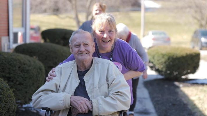 A smiling resident in a wheelchair with a staff member outdoors