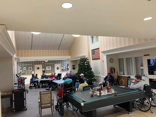 Group of seniors in wheelchairs seated around tables in a common area with holiday decorations.