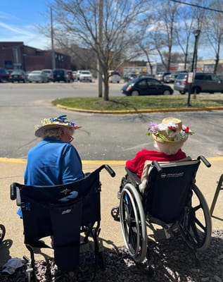 Two residents in wheelchairs wearing decorative hats, looking outdoors.