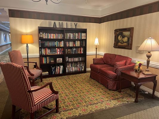 A cozy lounge with bookshelves, seating, and lamps in a senior living facility.