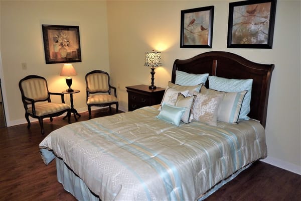 A well-decorated bedroom featuring a bed with light blue bedding, two armchairs, and framed artwork.