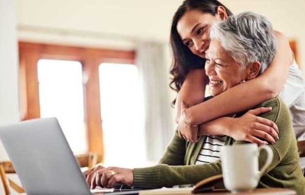 A senior woman smiles while using a laptop, embraced by a younger woman.