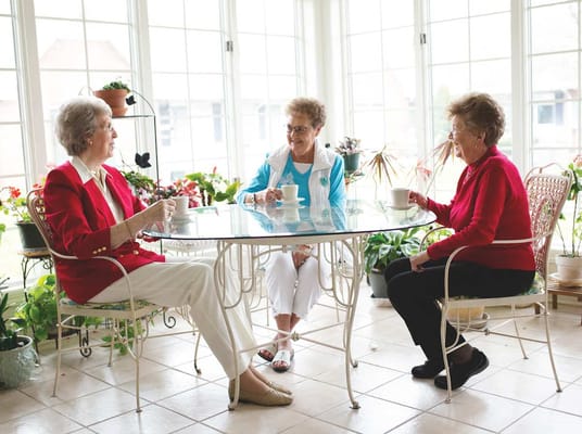 Three senior women seated at a table drinking tea surrounded by plants.