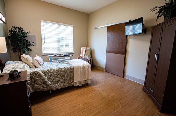 A cozy bedroom with a bed, chair, TV, and a small table in a senior living facility.