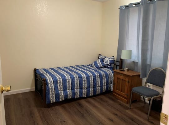 A cozy bedroom featuring a single bed, nightstand, lamp, and chair.