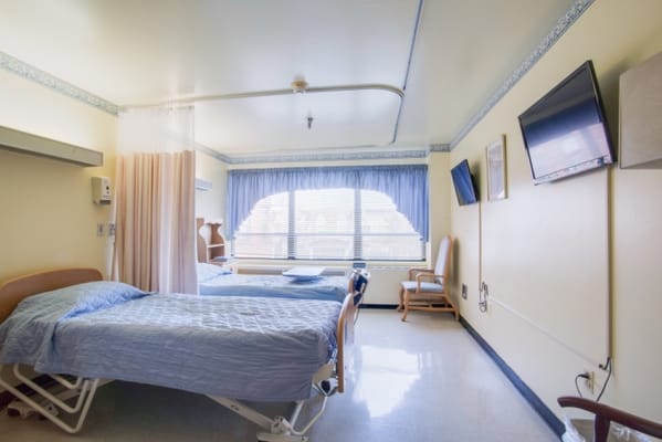 A shared bedroom in Stoddard Baptist Nursing Home with two beds and large windows.