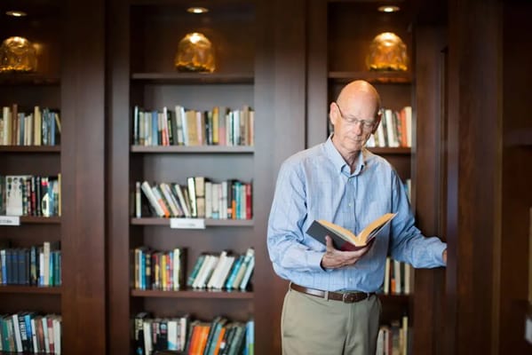 A senior man reading a book in a library filled with shelves of books.