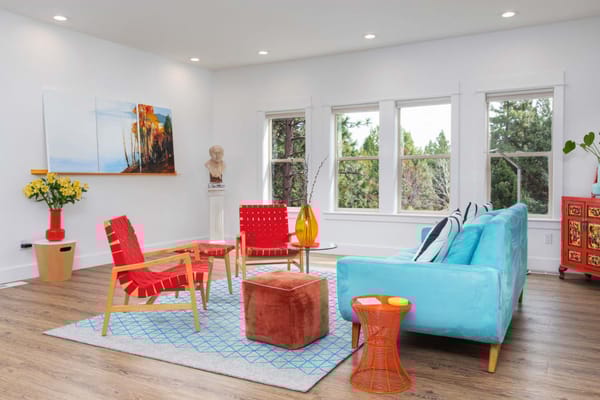 A colorful lounge with a blue couch, red chairs, and view of greenery through windows.