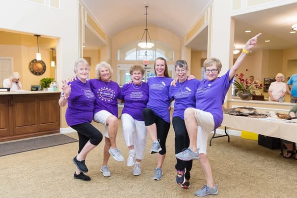 A group of six women wearing purple shirts dancing in the lobby of Touchmark on West Prospect.