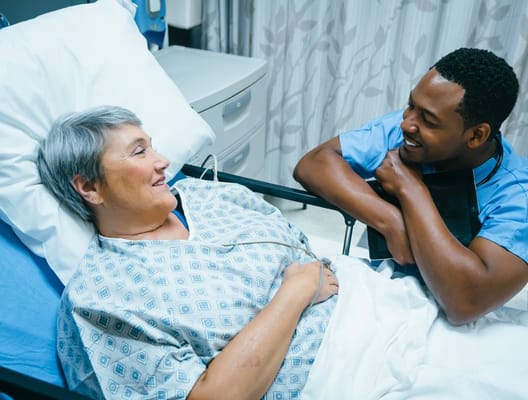 A caregiver smiles and chats with a patient in a hospital bed.