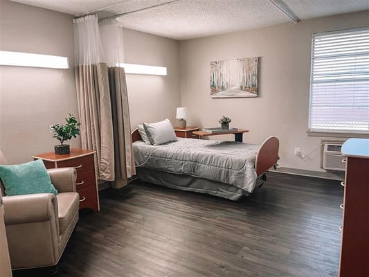 A well-lit resident room featuring a bed, chair, and small desk.