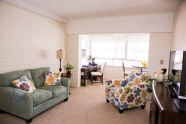 A cozy living room featuring a green couch, floral armchair, and a desk by the window.
