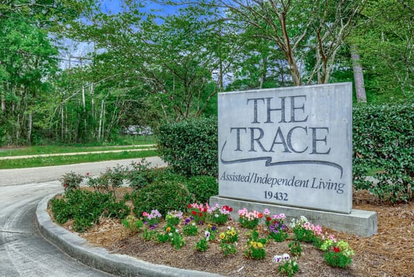 Sign for The Trace assisted living facility surrounded by greenery and flowers.