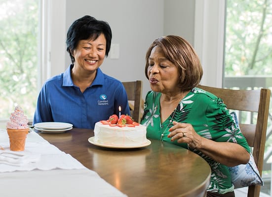 An older woman blowing out candles on a birthday cake while a caregiver smiles.