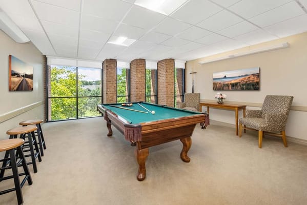 Billiards table in a well-lit room with large windows and seating areas.