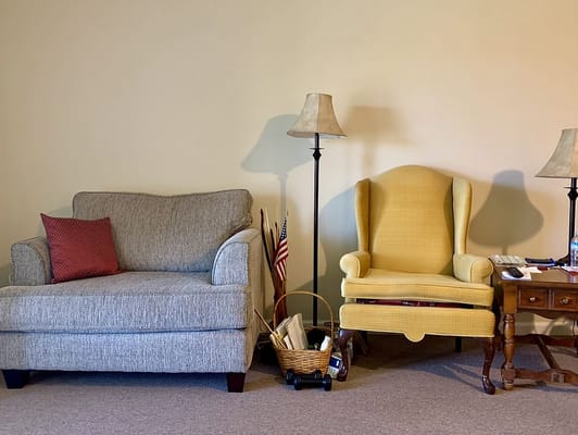 A gray loveseat and a yellow wingback chair beside a lamp and a small table