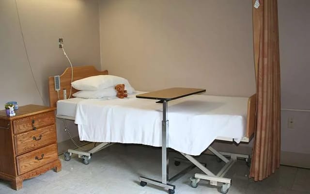 A hospital bed in a private room with a teddy bear on the pillow.