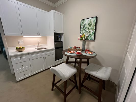 A kitchenette with a small dining table, two stools, and a bowl of fruits.