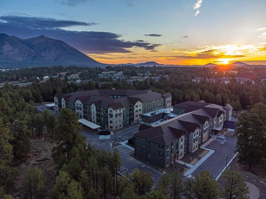 Aerial view of The Bluffs of Flagstaff Senior Living surrounded by pine trees and mountains at sunset.