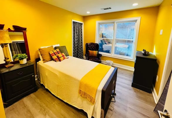 A cozy, well-lit bedroom with a yellow wall, a bed, and a vintage chair.