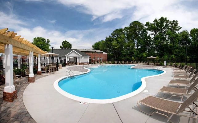 Swimming pool surrounded by lounge chairs with a pergola and trees in the background.