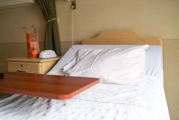 A neatly made bed with a wooden bedside table and a bag of oranges.