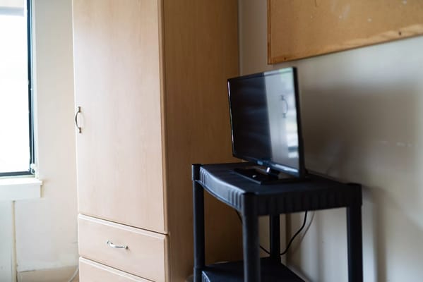 A television on a black stand next to a wooden wardrobe in a resident room.