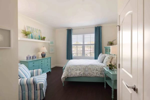 A well-decorated bedroom with a turquoise dresser, comfortable bed, and large windows.