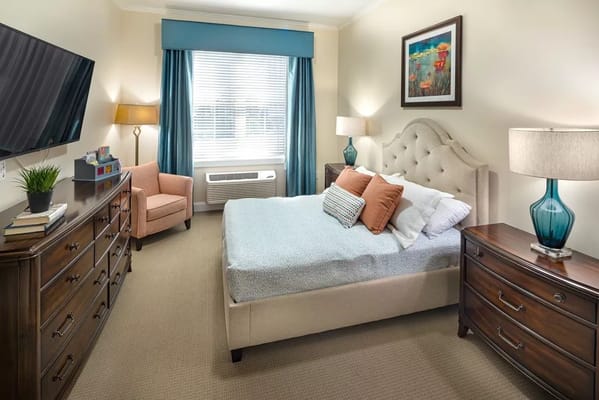 Cozy bedroom featuring a bed, dresser, and seating area in a senior living facility.