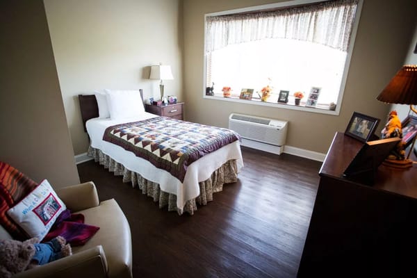A well-decorated bedroom in a senior living facility featuring a bed, nightstand, and window with natural light.