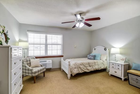 A cozy bedroom featuring a white bed, dresser, plaid chair, and window.