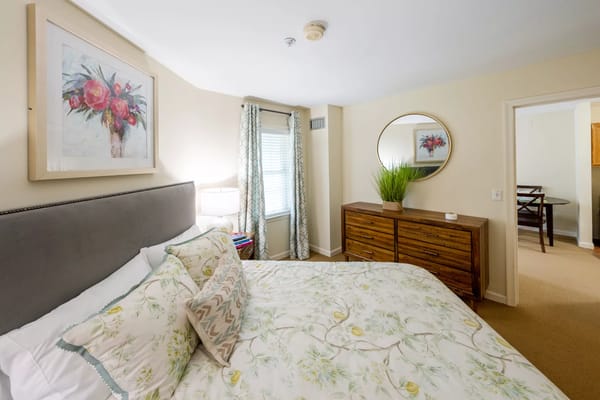 A bright and inviting bedroom featuring a bed with floral bedding, a wooden dresser, and large windows with curtains.