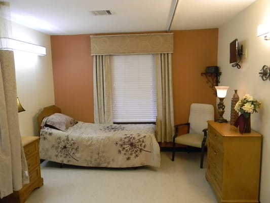 A cozy bedroom featuring a bed, chair, lamp, and dresser.