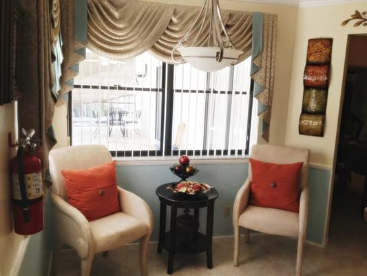 Two cream-colored chairs with orange pillows next to a small table with fruit, near a window with vertical blinds.